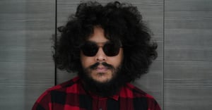 Young man with dark curly hair and sunglasses posing in a dark red shirt