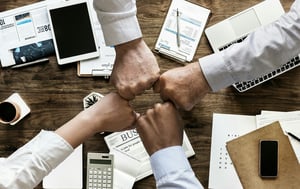 four fists bumping each other above a work desk filled with papers and laptops